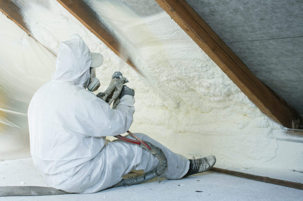 A worker in protective gear applying spray foam insulation to an attic, illustrating the process of improving home energy efficiency.